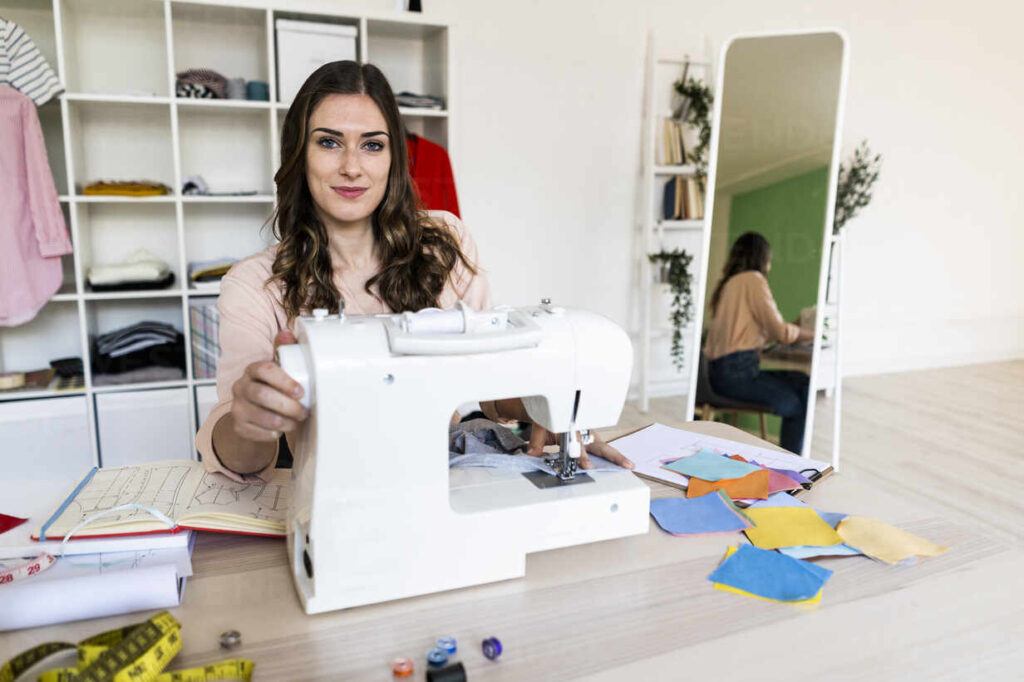 smiling-female-creative-tailor-sitting-with-sewing-on-machine-at-studio-GIOF09808